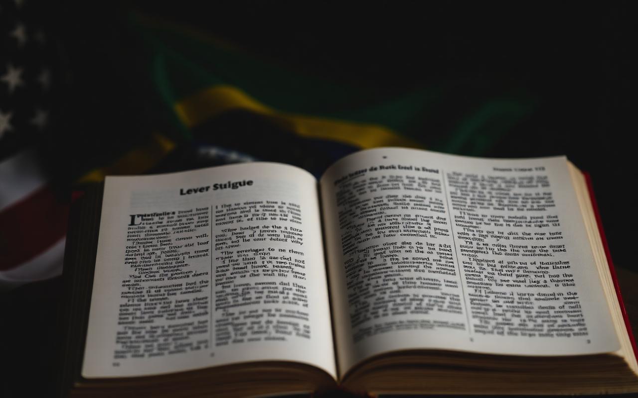 Livro aberto sobre mesa escura com bandeiras do Brasil e dos Estados Unidos desfocadas ao fundo, simbolizando tradução.